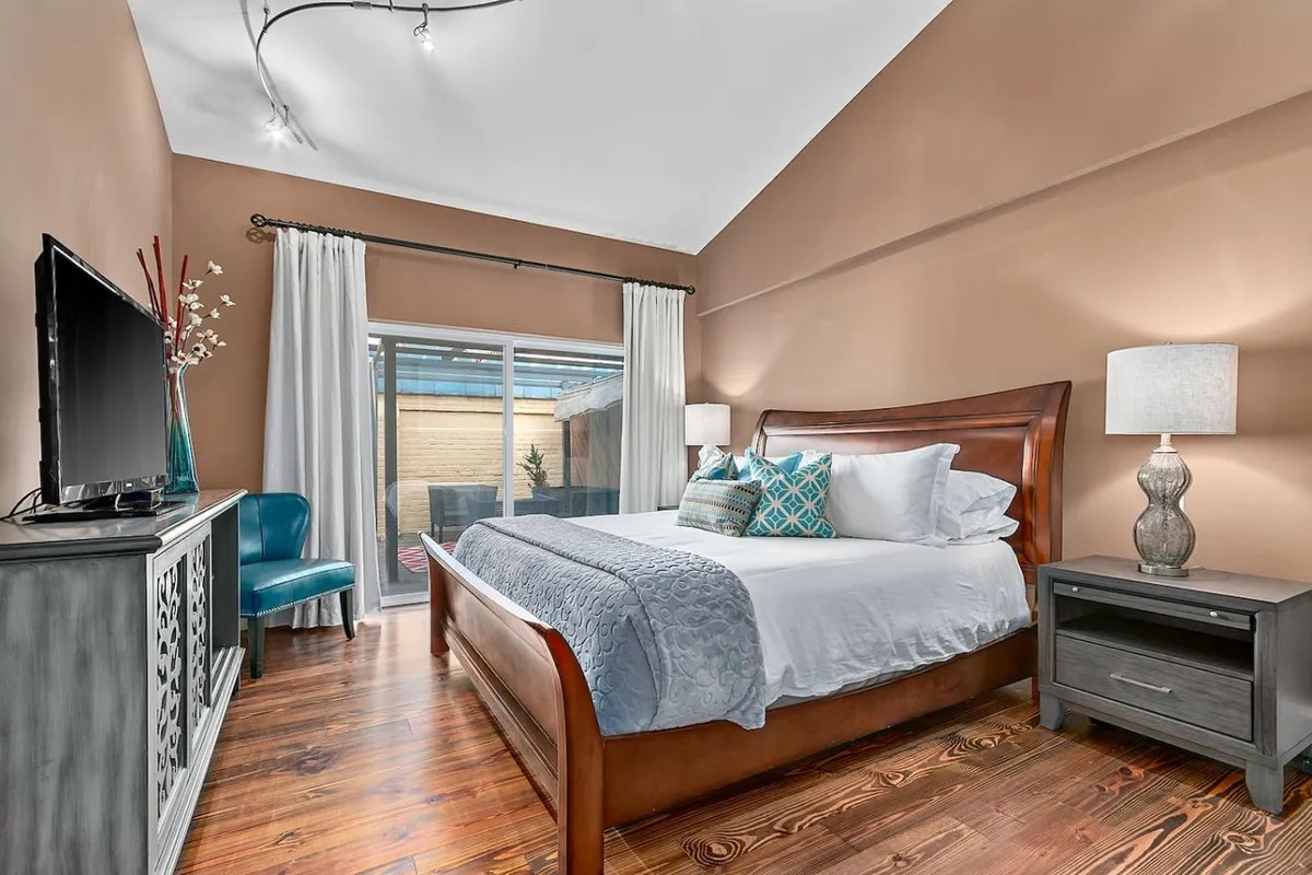 A serene and cozy bedroom scene featuring a comfortable bed with decorative pillows, a modern lamp, TV, and wooden furniture. Sunlight streams in, suggesting both relaxation and the autonomy of personal space above city life.