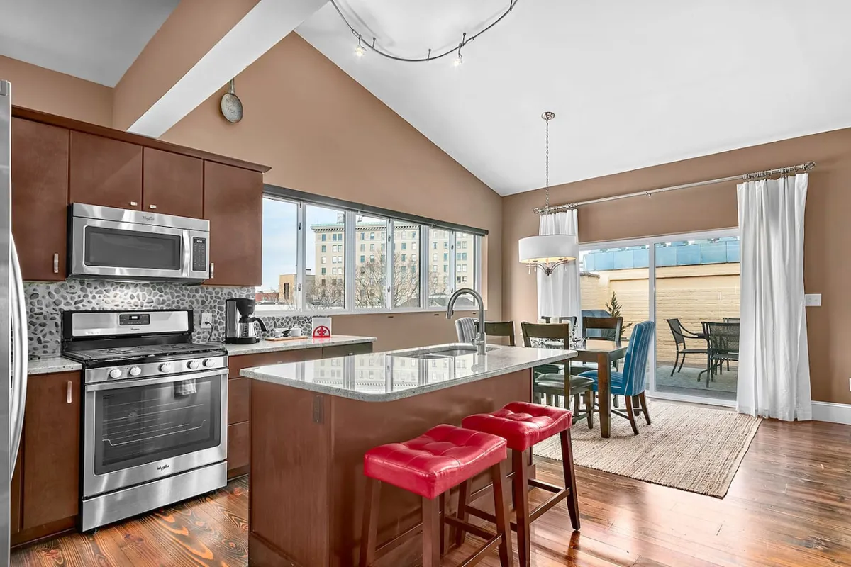 A bright, welcoming dining and kitchen area featuring a kitchen island, stainless steel appliances, spacious dining table, and expansive windows letting in daylight. The room feels ready for entertaining, brunches, or festive dinners.
