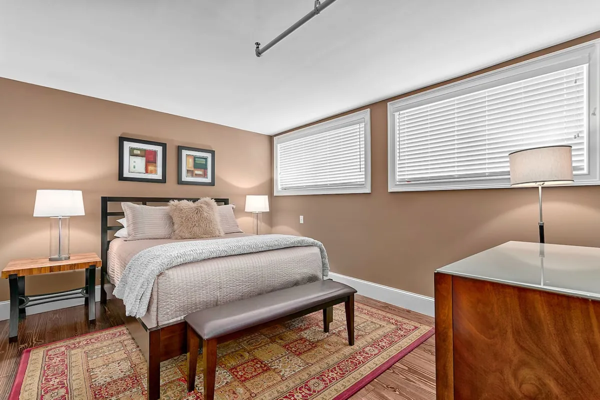 A spacious and tranquil bedroom with a plush bed, coordinating nightstands, elegant wall art, a soft bench at the foot of the bed, and a glowing lamp. Sunlight streams through the windows, enhancing the room's peaceful mood.