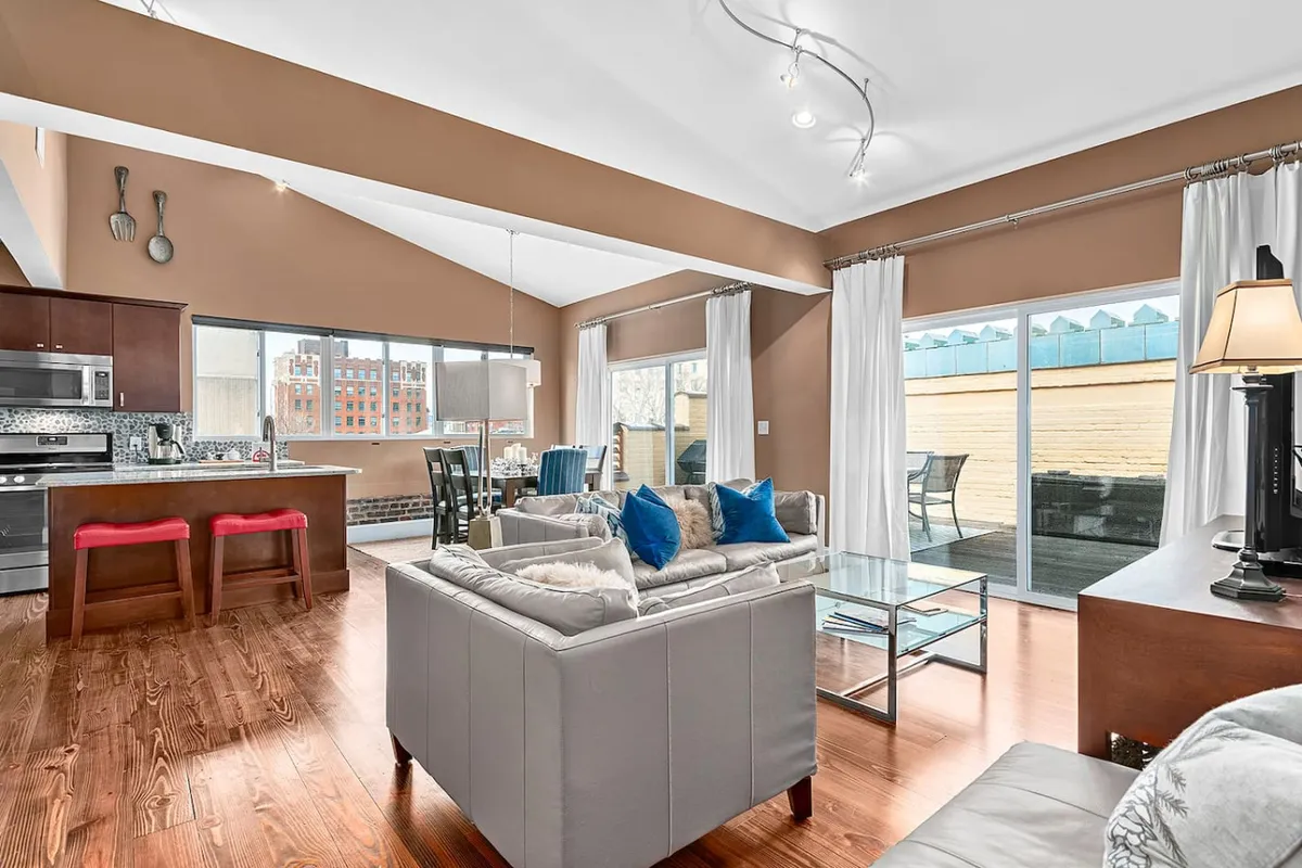 A warm, welcoming open-plan living space with a modern sofa, dining table, and kitchen island, all set against a backdrop of large windows that flood the room with natural light. The furnishings reflect Art Deco-inspired lines and materials, and the setup invites socializing, relaxing, and enjoying meals together.