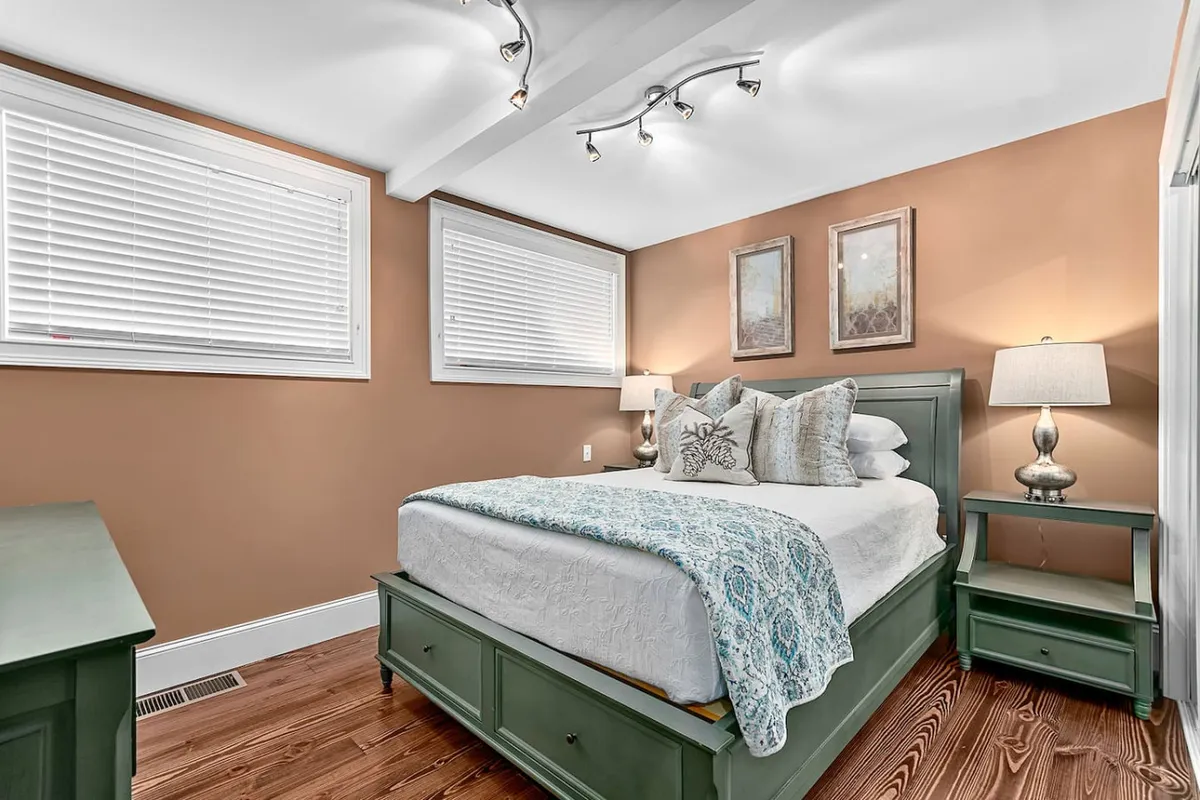 A tranquil bedroom featuring a comfortable bed adorned with decorative pillows, a soft throw, and a green dresser. Natural light gently illuminates the room, and a cozy table lamp and framed artwork complete the relaxing atmosphere.