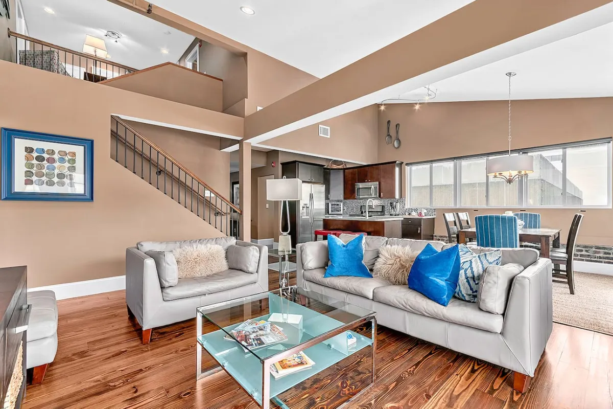 The image showcases a spacious, stylish living area with plush gray sofas adorned with decorative pillows, a sleek glass coffee table, and an open-concept kitchen in the background. A modern staircase leads to the upper level, hinting at the apartment's generous layout. The scene exudes comfort and is perfectly set for gatherings, relaxing evenings, or socializing with friends and family.