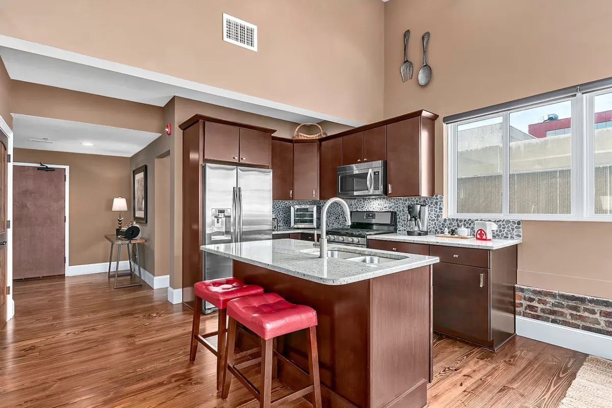 A contemporary kitchen featuring a spacious island with striking red stools, stainless steel appliances, and elegant granite countertops. Sunlight pours in through a large window, illuminating the space and creating a welcoming environment for cooking and entertaining.